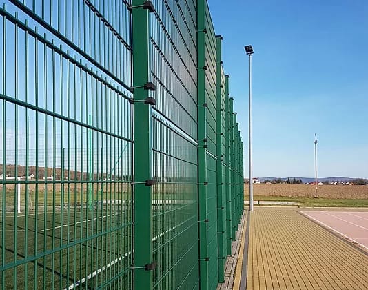 Green twin-wire mesh sports fencing running alongside an artificial pitch